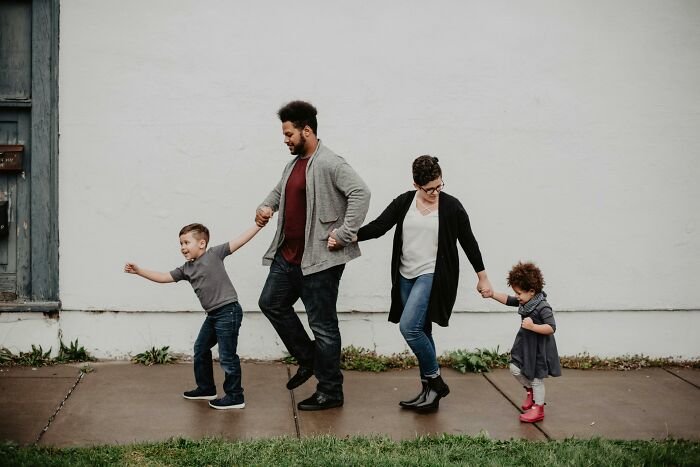 A family of four walking together outside, laughing and holding hands near a white wall.