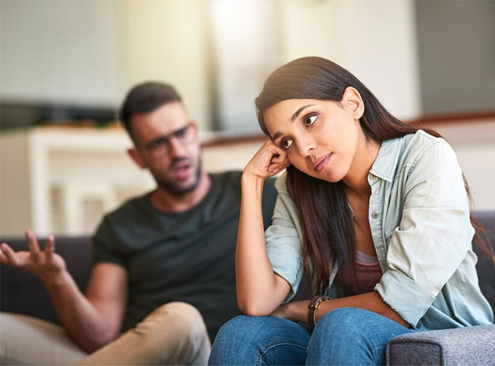 Couple arguing about Hispanic cooking, man gesturing and woman looking away, both seated in a living room.