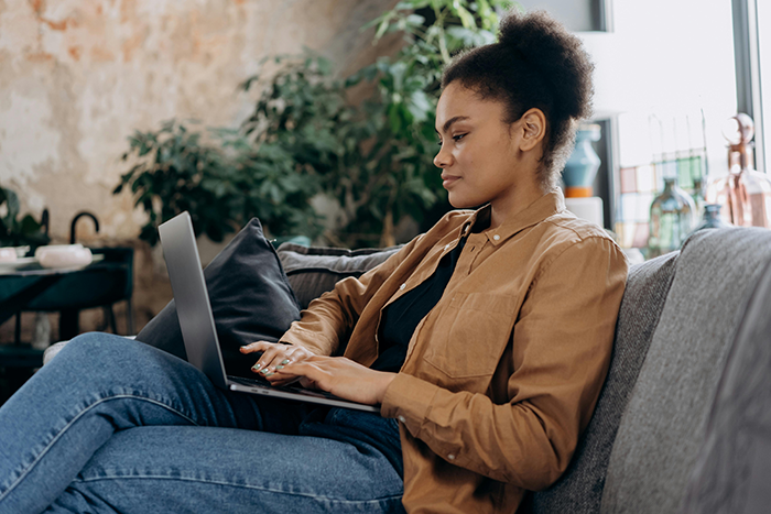 Woman on laptop, possibly planning a honeymoon, sitting on a couch in a cozy setting with plants.