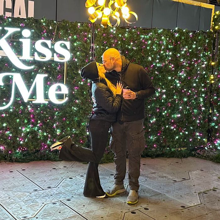 Couple kissing in front of "Kiss Me" sign with festive lights, wearing casual winter attire.