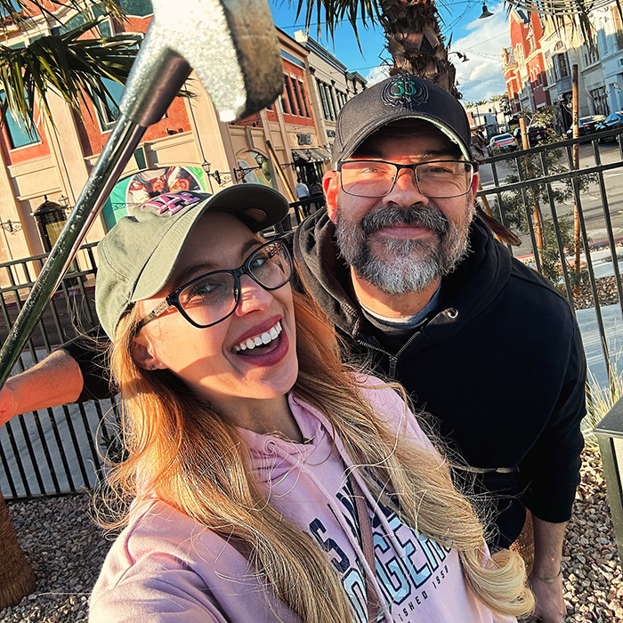 Smiling couple poses for a selfie, with greenery and buildings in the background.