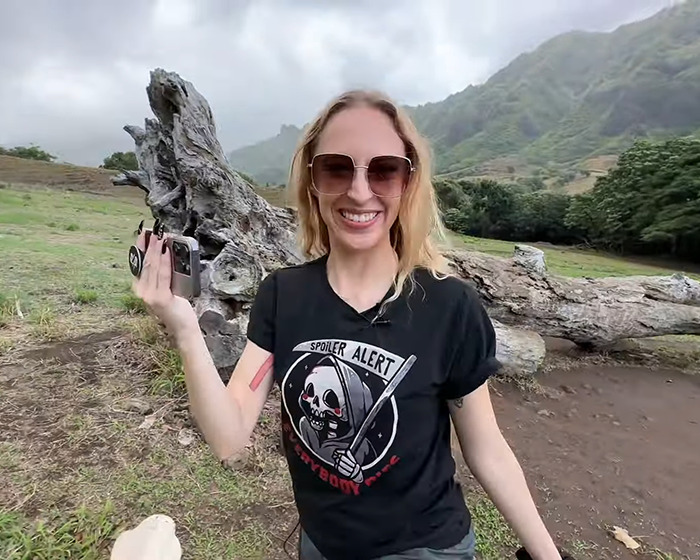 TV star&rsquo;s wife smiles sinisterly, holding a camera in a scenic outdoor setting.