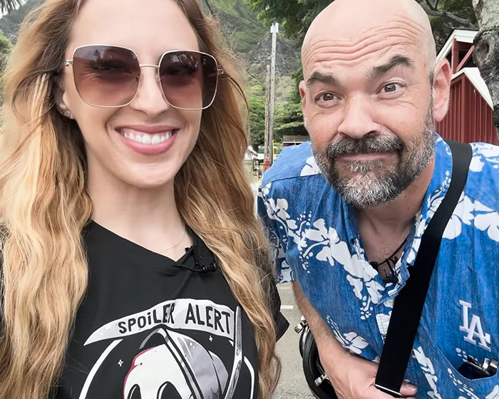 TV star's wife smiling sinisterly in sunglasses, standing outdoors with a man in a blue shirt.