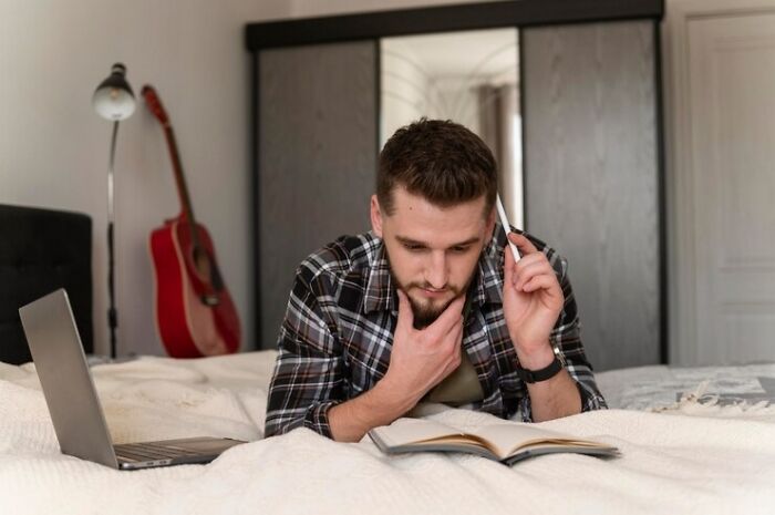 Man in a plaid shirt on a bed, reading a book, contemplating about a situation involving a spare room and an unexpected guest.