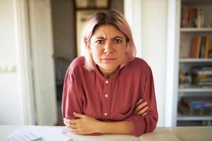 Person in pink shirt looking upset while seated at a table, implying tension in a shared living situation.