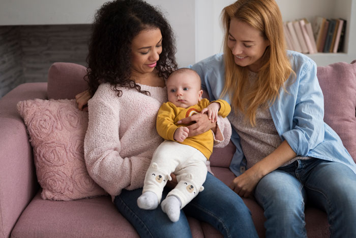 Two women smiling at a baby sitting on their lap, celebrating a friend's birthday together on a pink sofa.