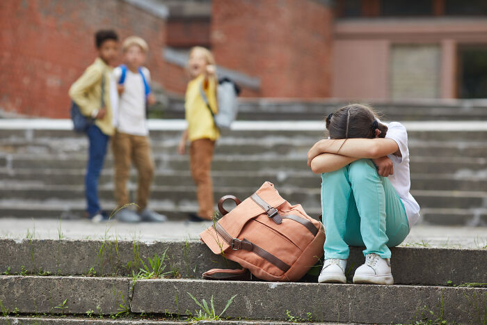 Child sitting alone on steps looking sad, with backpack beside them, while other kids in background talk, depicting insignificant thing.