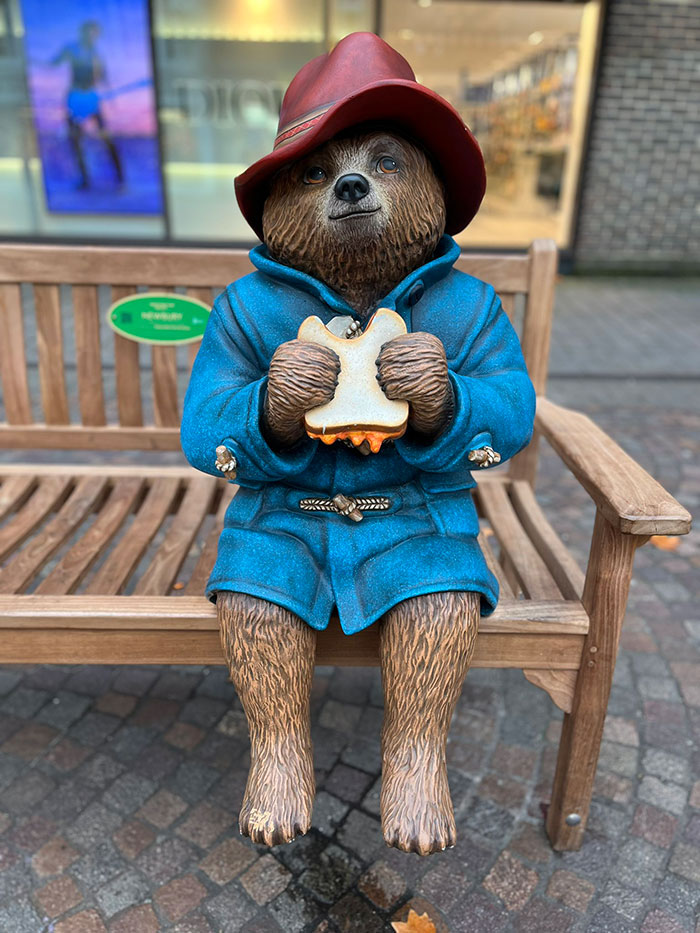 Paddington Bear statue in a blue coat and red hat, sitting on a bench, holding a sandwich.