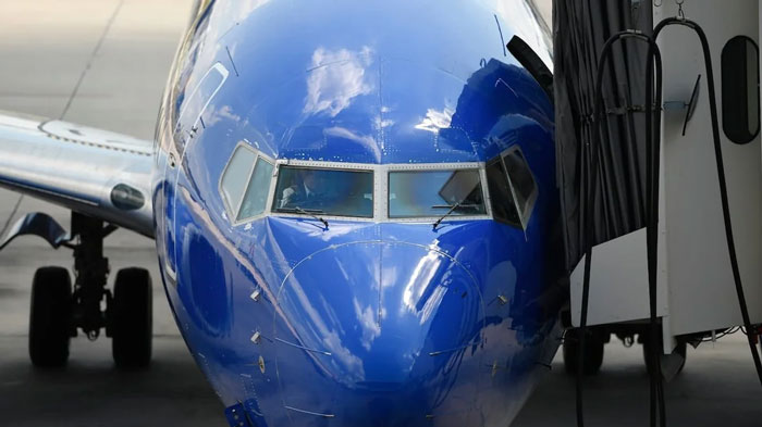 A blue airplane nose docked at an airport gate, reflecting clouds, related to Florida Man headlines.