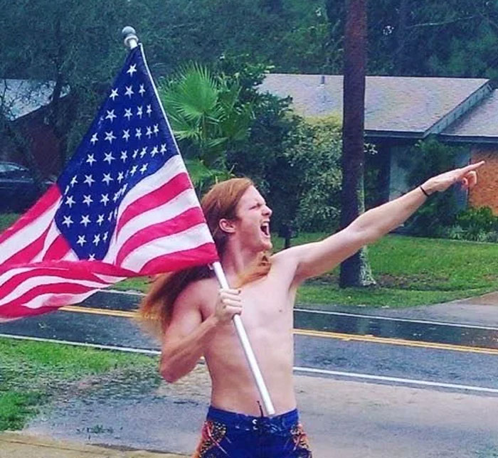 A shirtless man with long hair holds an American flag and points outward in a dramatic pose, embodying a Florida Man moment.