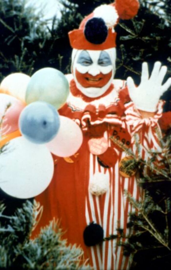 Creepy clown in red and white costume holding balloons, surrounded by trees.