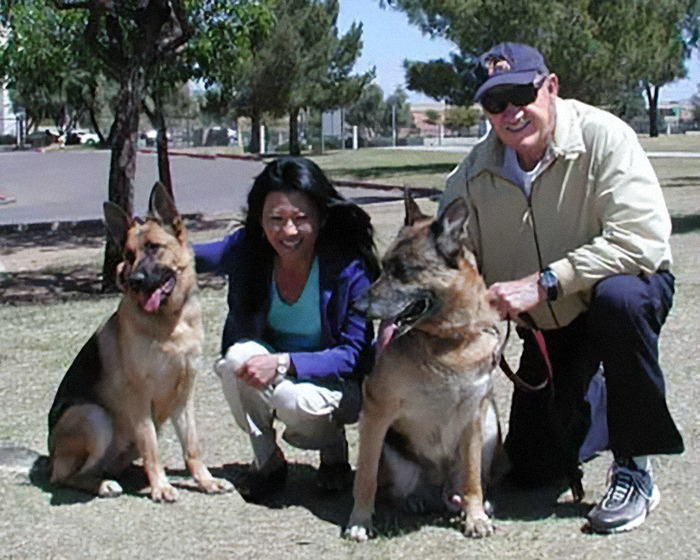 Gene Hackman and Betsy Arakawa smiling with two German Shepherds at a park.