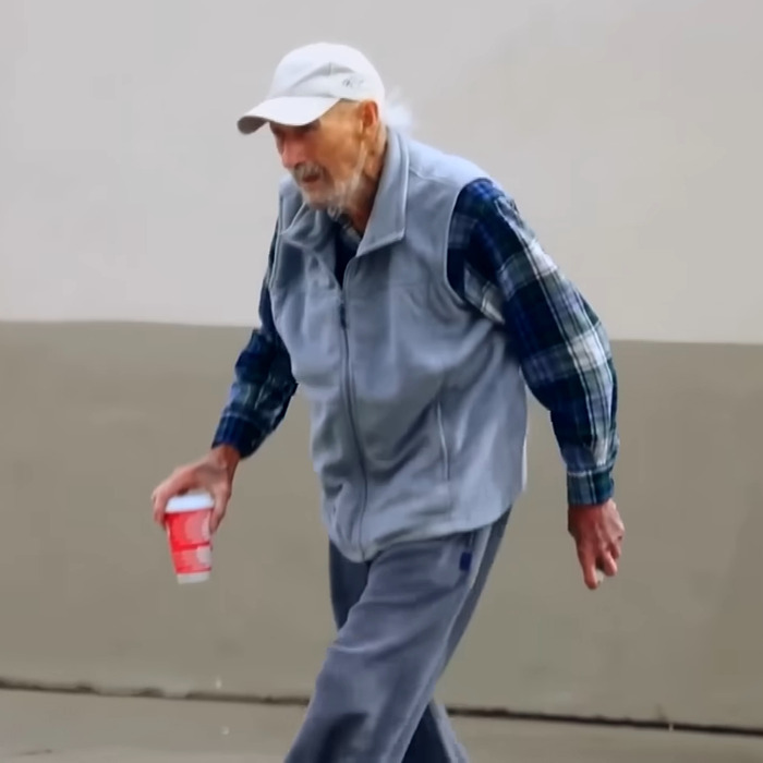 Gene Hackman in casual attire holding a cup, walking outdoors.