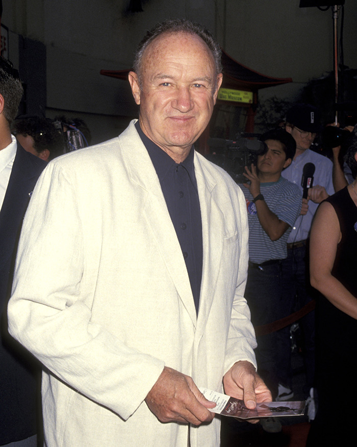 Gene Hackman in a white jacket at an event, holding a paper, surrounded by people and cameras.