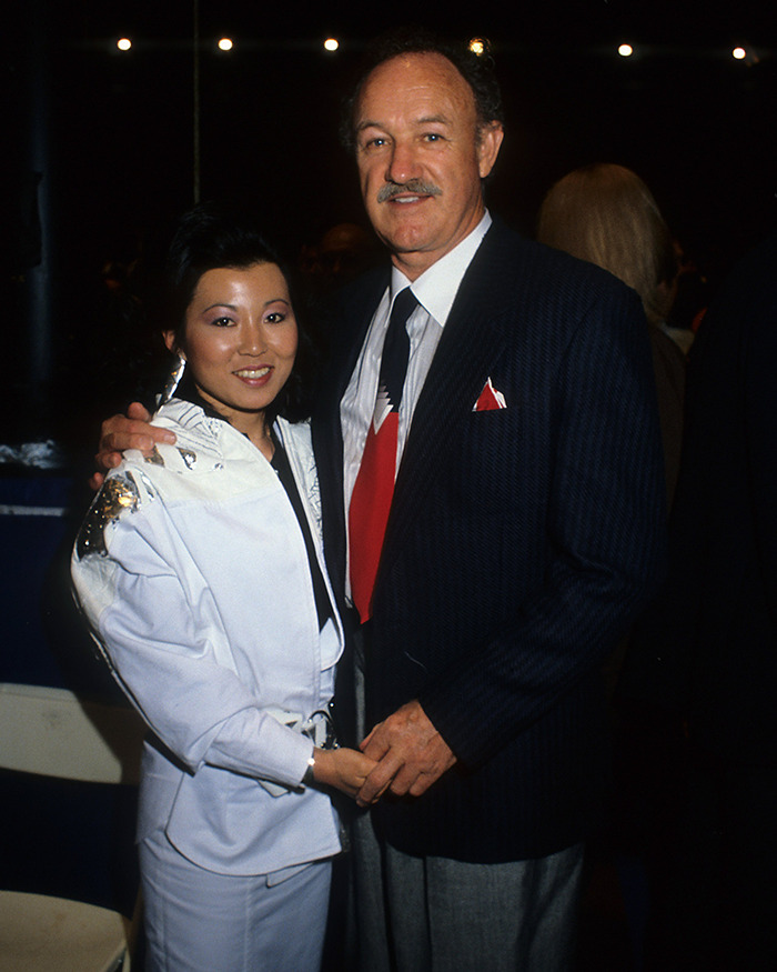 Gene Hackman and Betsy Arakawa posing at an event, man in suit and woman in a white outfit, both smiling.