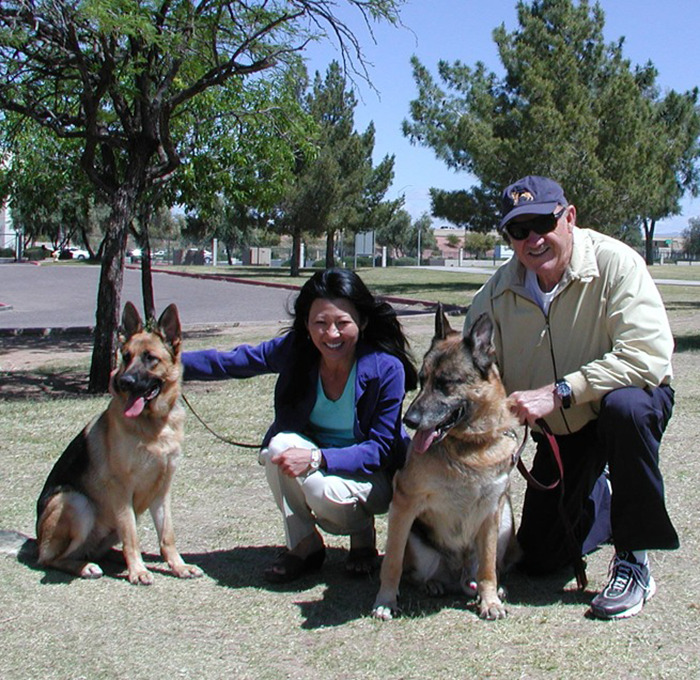 Man and woman with two German Shepherds in a park, smiling under trees, related to Gene Hackman's family and results issue.