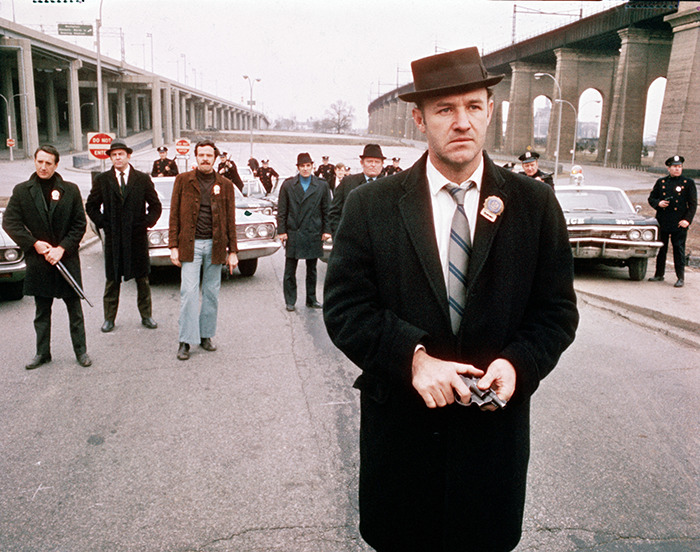 Gene Hackman in a classic film scene wearing a hat and coat, looking serious, with vintage police cars behind him.