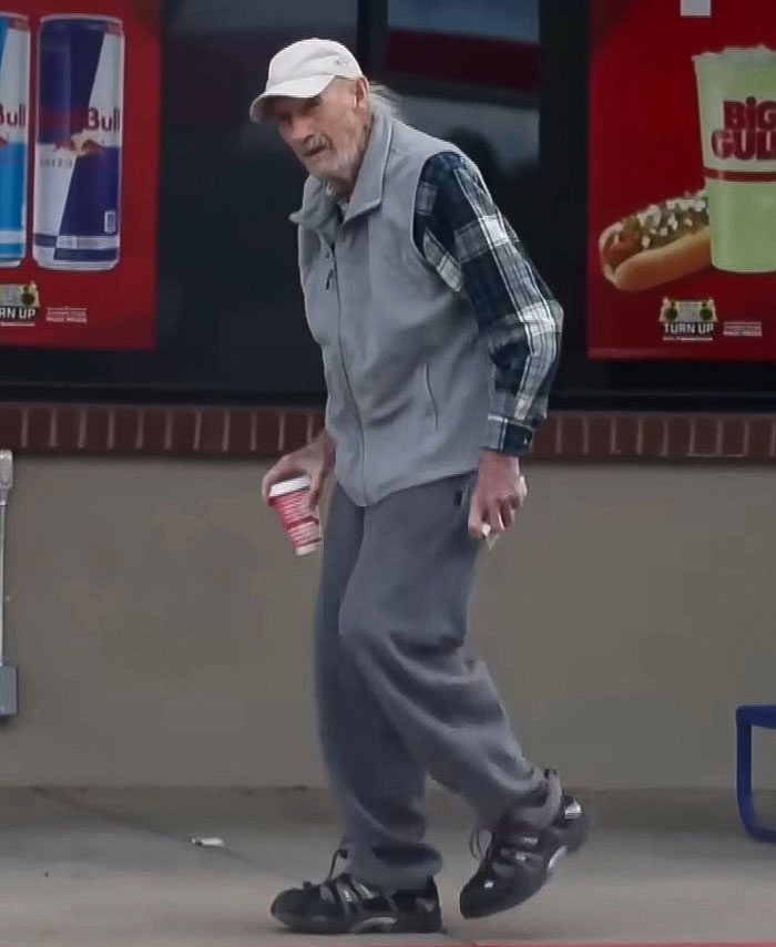 Man in casual outfit holding a cup, standing outside a store with signage in the background.