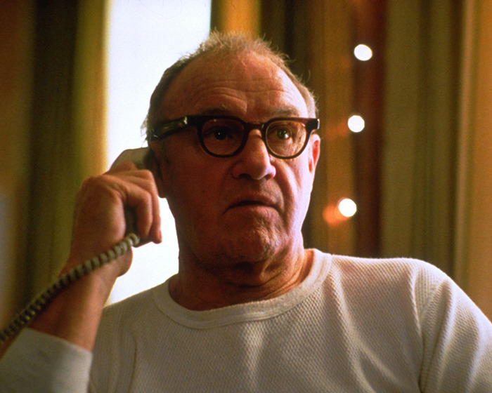 Gene Hackman wearing glasses and white long sleeve while talking to the phone indoors in a movie scene.