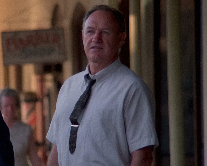 Gene Hackman on a street, wearing a tie and shirt, looks into the distance with a blurred backdrop of a shop sign in a movie scene.