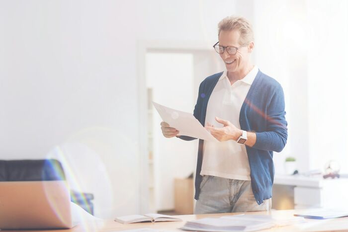 Man in glasses smiling, reading documents at a desk in a bright room, symbolizing personal secrets and family privacy.