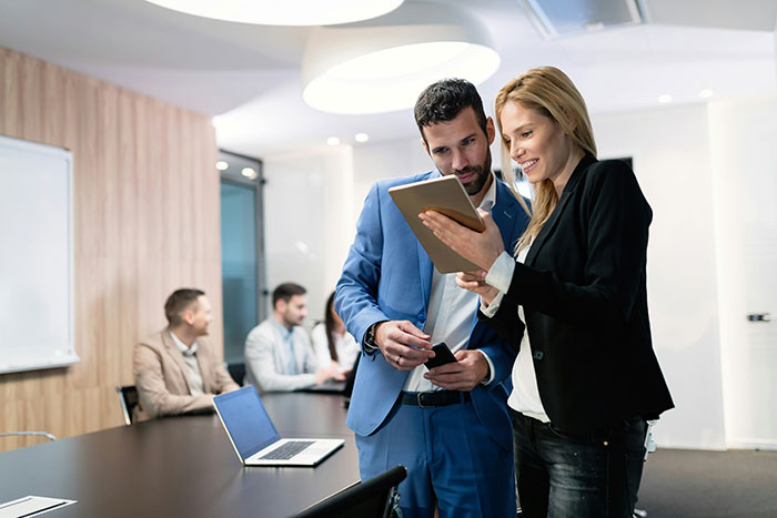 Two colleagues discussing a project in a modern office setting, with others working in the background.
