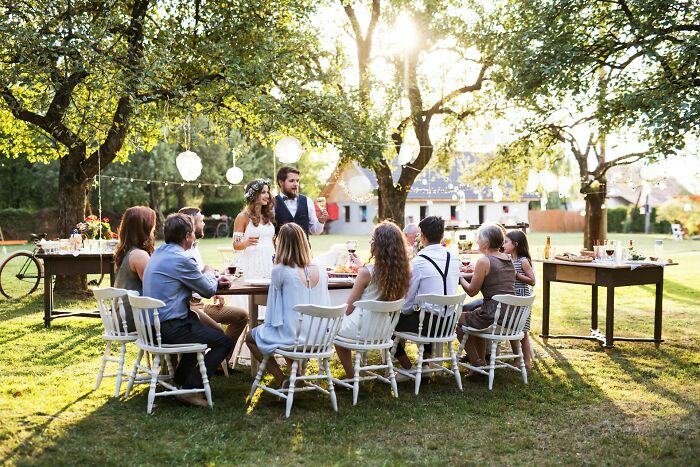 Outdoor wedding scene with people seated at a decorated table, celebrating under trees with hanging lanterns.