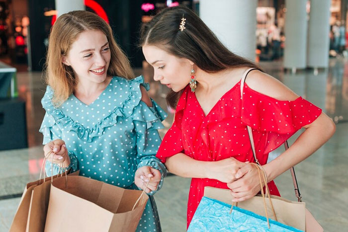 Two teenage girls wearing colorful dresses, smiling while shopping, holding bags in a mall.
