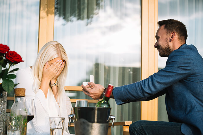 Man proposes with a ring in a box to a woman covering her face at a table with wine and roses.