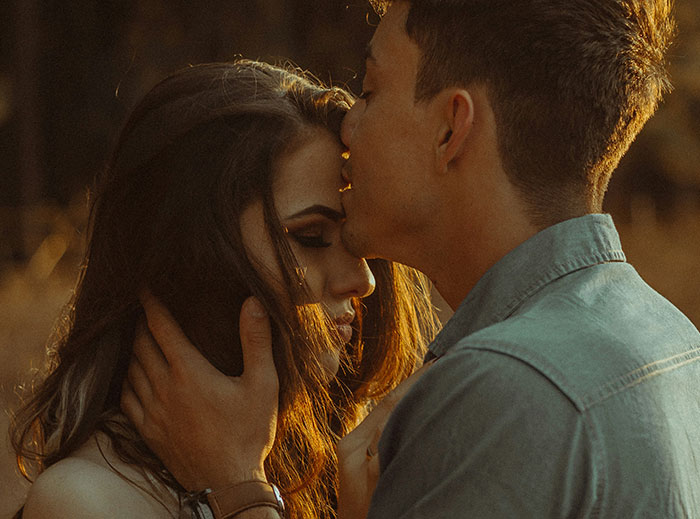 A man gently kisses a woman's forehead in a serene outdoor setting, exemplifying a "nice guy" moment.