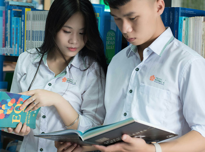 Two students in a library, both in white uniforms, reading together with focused expressions, showcasing academic engagement.