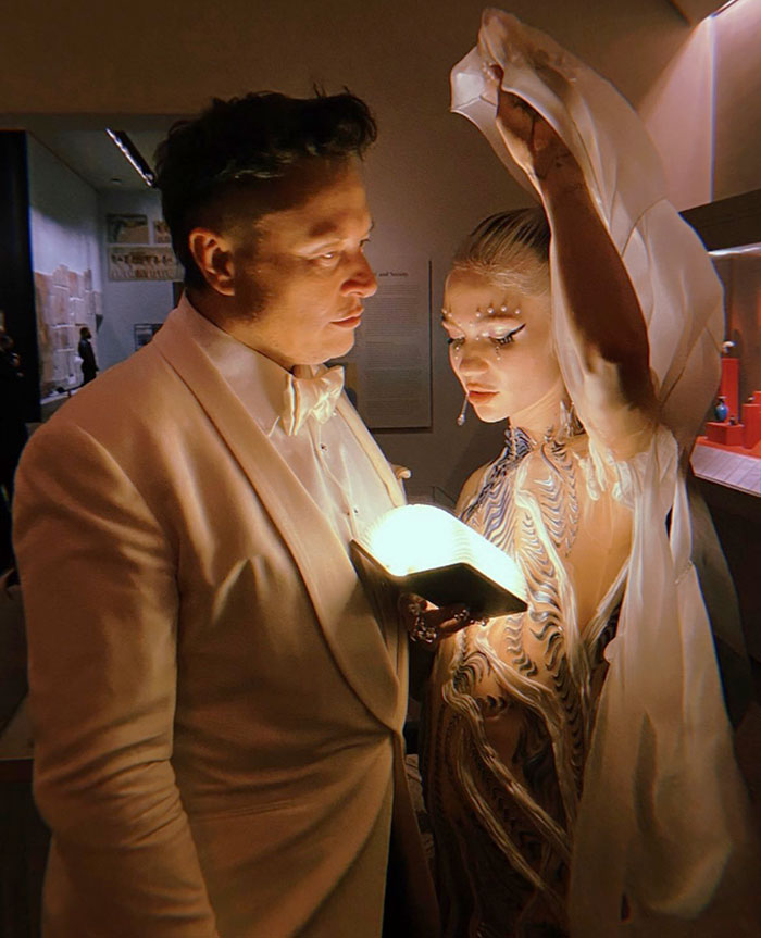 Man in white jacket and woman in detailed dress under low light, discussing while holding an illuminated book.