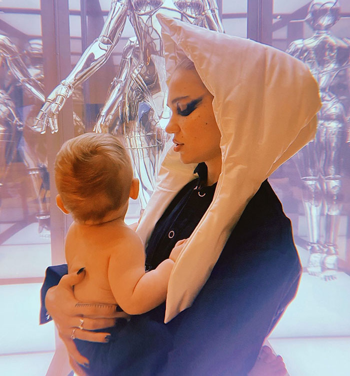 Child held by a person in a white hood, with reflective robotic figures in the background.