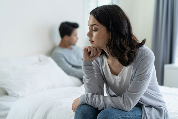 Couple sitting apart on bed; girlfriend looks upset, highlighting relationship tension. Couple sitting apart on bed; girlfriend looks upset, highlighting relationship tension.