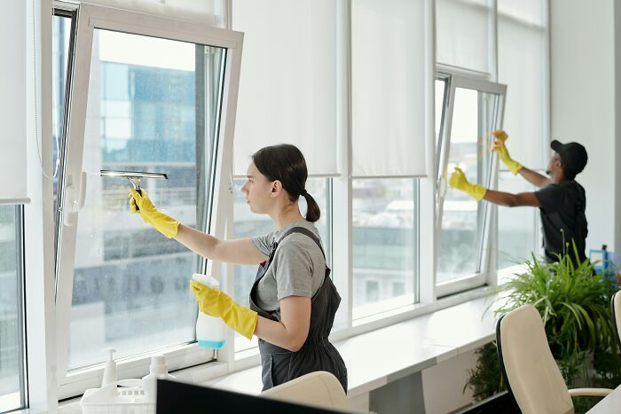 People using cleaning hacks to wash large windows, wearing yellow gloves and aprons in a bright room.