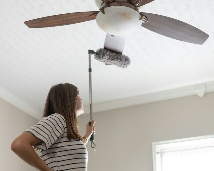 Woman using cleaning hack to dust ceiling fan with extendable tool.