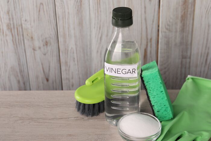 Vinegar bottle, brush, sponge, and gloves for cleaning hacks on a wooden surface.