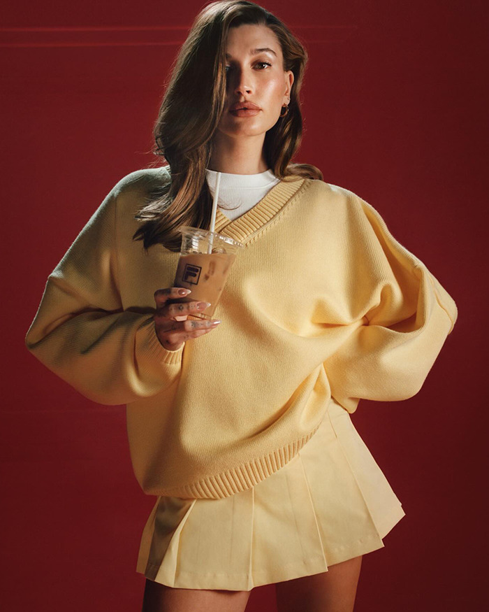 Hailey Bieber in yellow sweater holding iced coffee, posing against red backdrop.