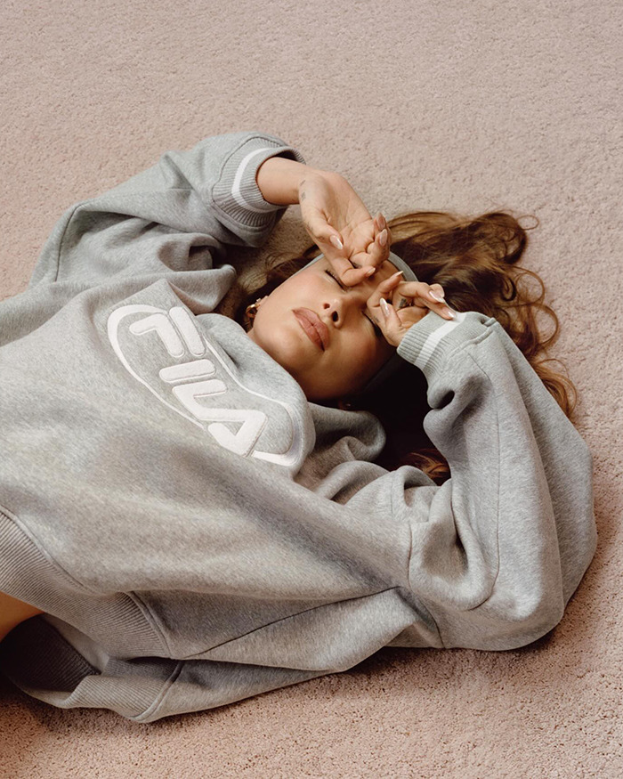 Hailey Bieber in gray sweatshirt lying on the floor, hands over face.