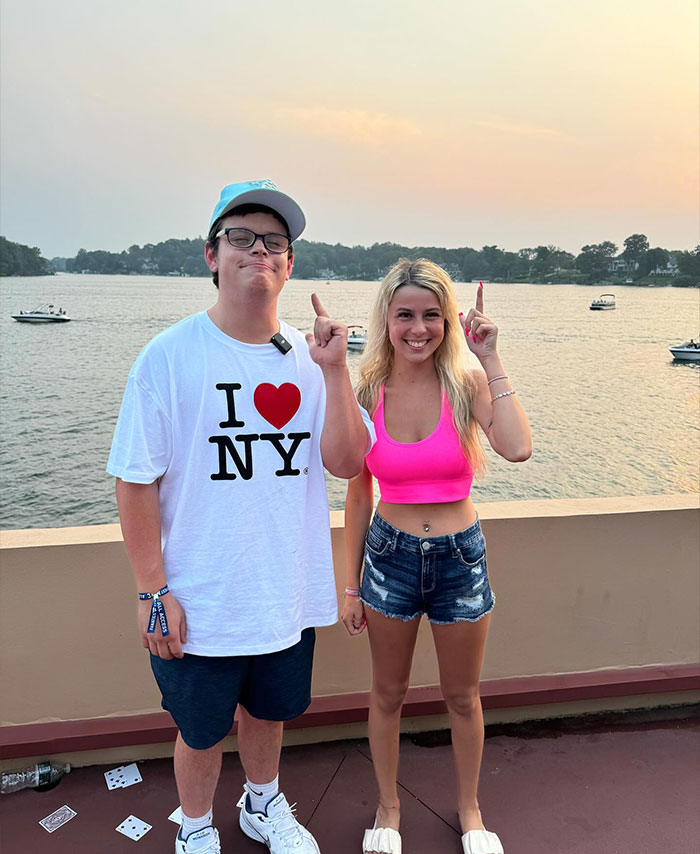 Two smiling individuals, including Hailey Welch, on a lakeside balcony, enjoying the sunset, wearing casual summer attire.
