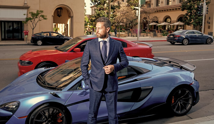 Man in a suit standing next to a luxury car on a city street, highlighting what society accepts as normal.