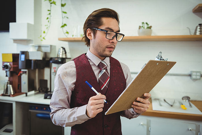 Man in glasses and vest holding a clipboard, assessing kitchen operations in a restaurant setting.