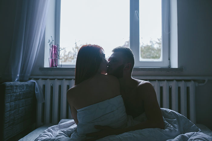 A couple kissing in bed, wrapped in blankets, with light streaming through a window.