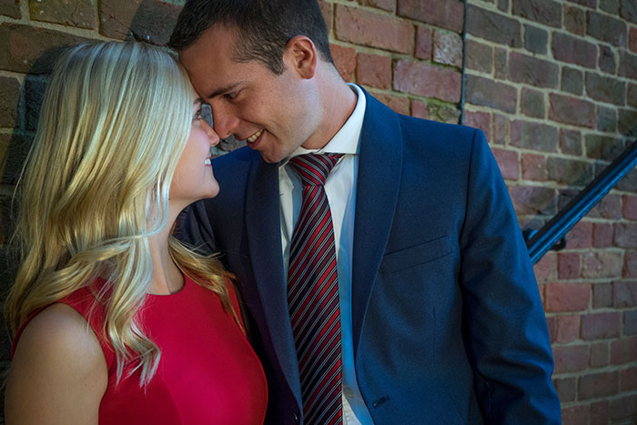 A couple in formal attire smiling intimately, hinting at more than just friendship, near a brick wall.