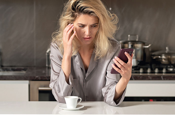 A woman looks concerned while reading a message on her phone, referencing "just a friend" lies, in a kitchen setting.