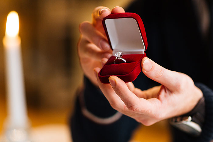 Hands presenting an engagement ring in a red box, symbolizing deceptive relationship secrets.