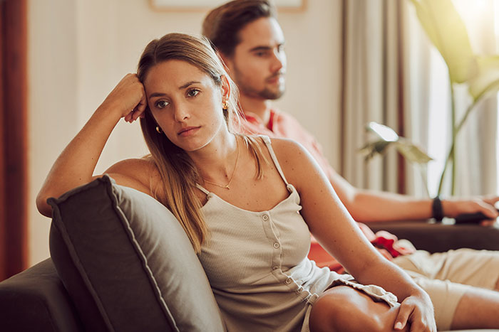 Woman looking concerned on a sofa with a man in the background, suggesting tension beyond "just a friend."