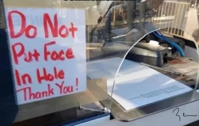 Humorous warning sign reading "Do Not Put Face In Hole" on a glass barrier.
