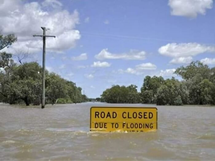 Flooding aftermath with road closed sign submerged in water under a cloudy sky.