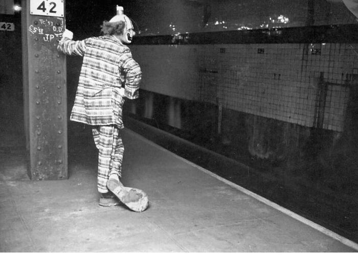 Clown in plaid costume leaning against a subway pillar, waiting on an empty platform, from forgotten corners of history.